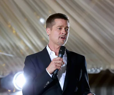 A person in a suit speaks into a microphone at an event with a well-lit backdrop and overhead drapery.