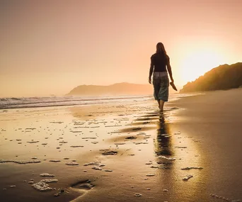 A person walking barefoot on a beach at sunset, leaving footprints in the sand.