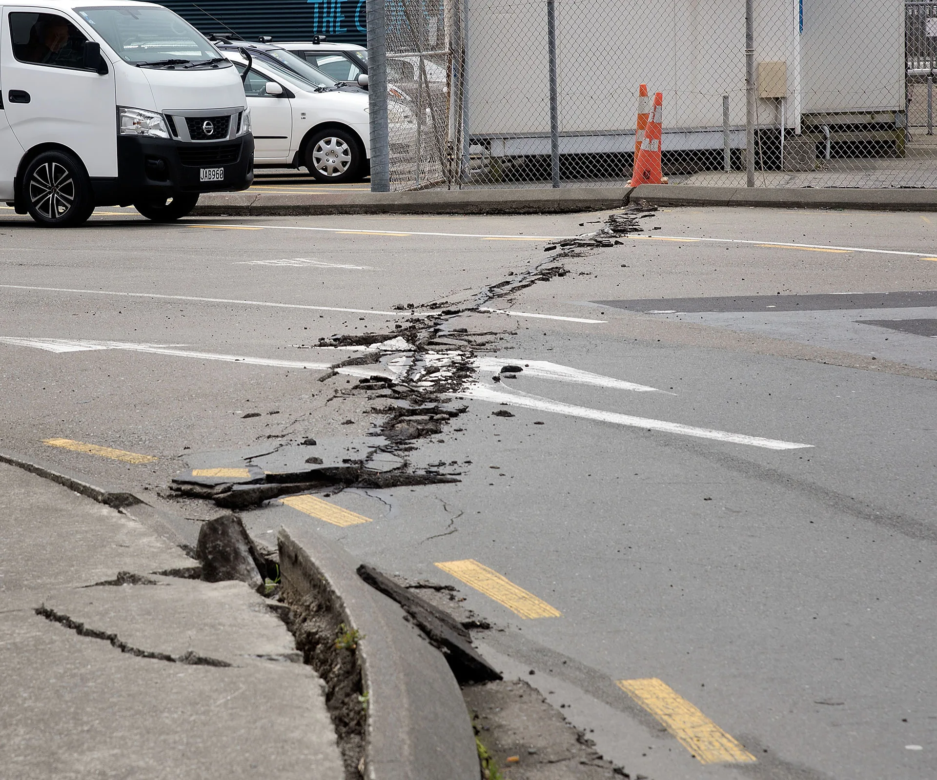 Cracked road with visible earthquake damage, debris scattered, and a white van nearby.