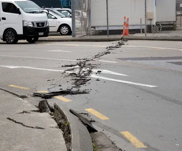 Cracked road with visible earthquake damage, debris scattered, and a white van nearby.