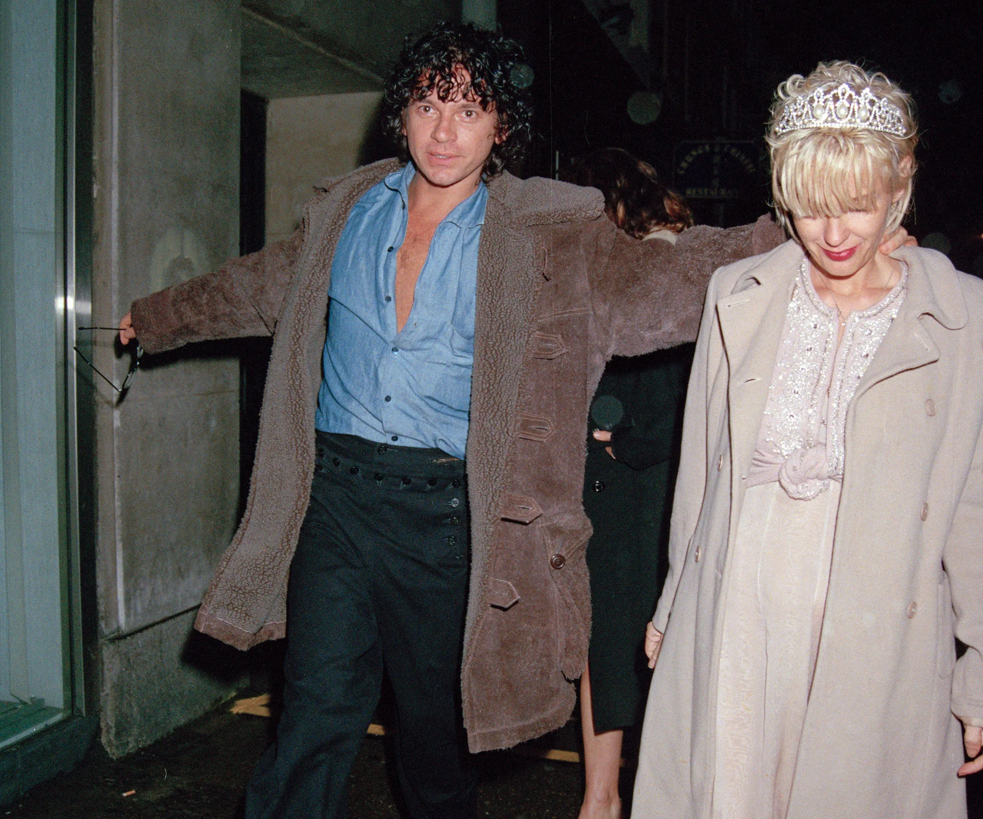 A person in a coat and another in a tiara walk together at night outside a building.