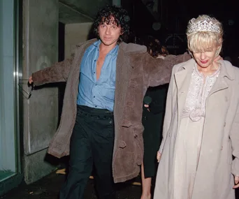 A person in a coat and another in a tiara walk together at night outside a building.