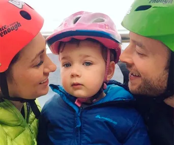 A child wearing a pink helmet stands between two adults in colorful helmets, all smiling.