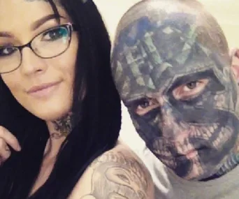 Tattooed woman with glasses and man with full facial tattoos posing for a selfie.