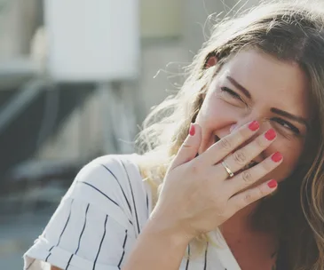 Smiling woman with red nail polish covering her mouth, wearing a striped shirt.