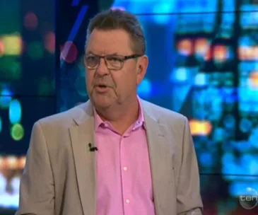 A man in glasses and a beige jacket over a pink shirt speaks on a TV show with a blurry, colorful background.