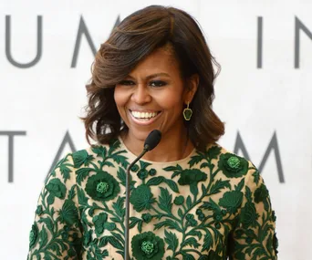 Michelle Obama smiling, wearing a green embroidered dress, speaking at a podium.