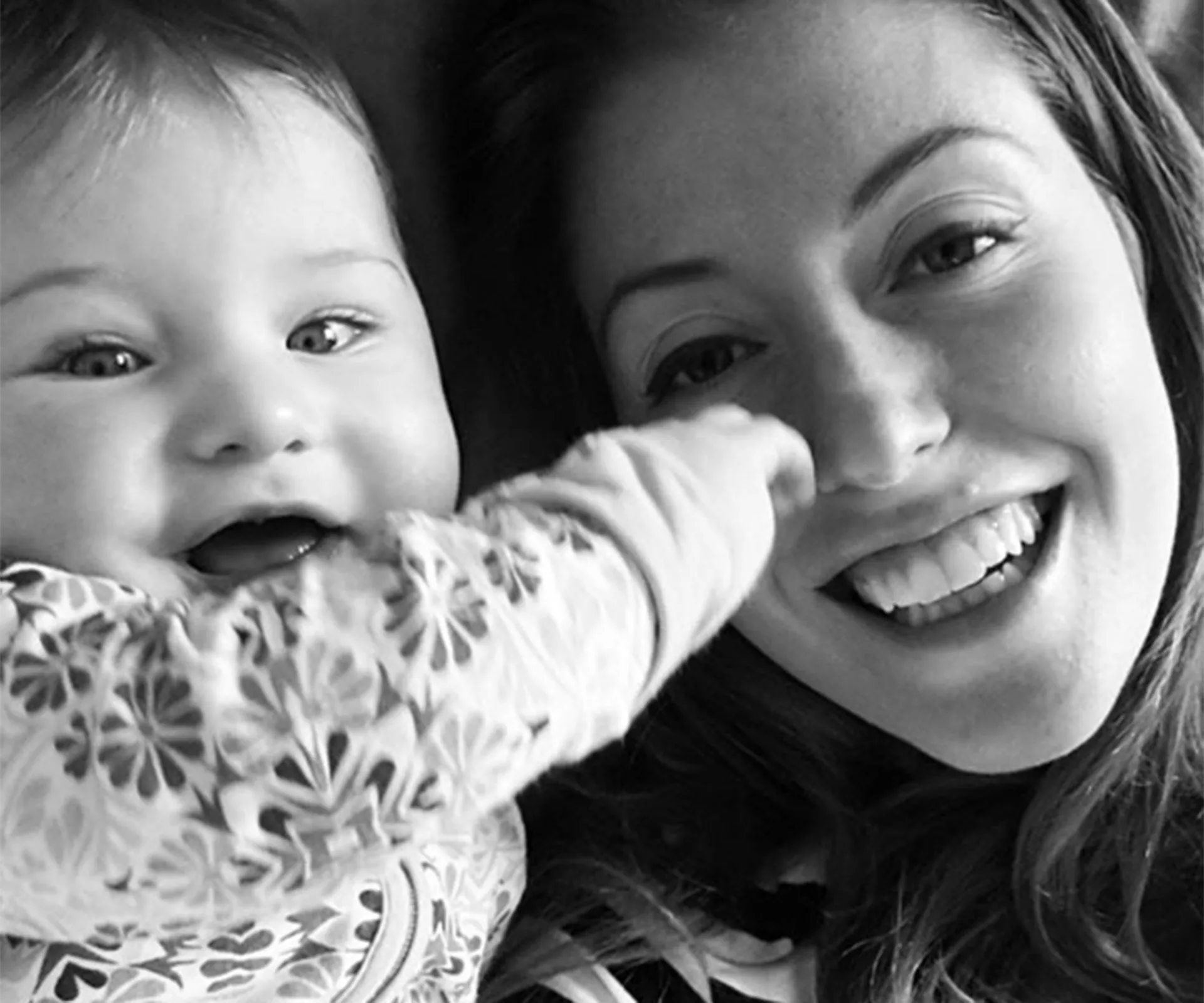 Mother and baby smiling together, with baby's hand touching mother's face, in a joyful black-and-white photo.