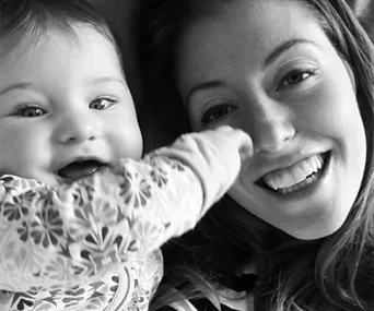 Mother and baby smiling together, with baby's hand touching mother's face, in a joyful black-and-white photo.