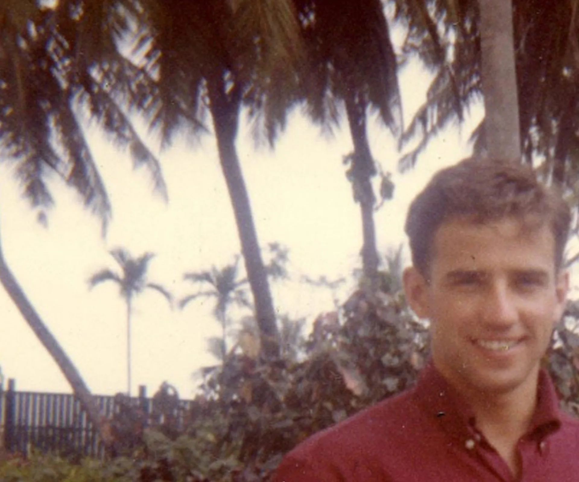 A young man in a red shirt smiles outdoors with palm trees in the background.