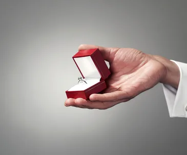 Man holding open red ring box with diamond engagement ring inside, against a neutral background.