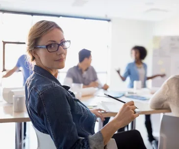 A woman with glasses holding a pencil looks back during a meeting in a bright conference room.