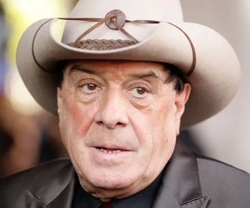 Man in a hat looking serious, wearing a dark suit, appearing outdoors.