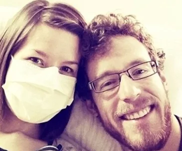 Woman wearing a mask sitting beside a smiling man with glasses, both appearing happy.