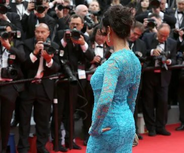 A woman in a blue lace dress poses on the red carpet as photographers gather around, capturing the moment.