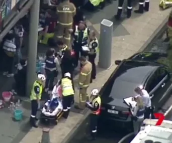 Emergency responders are attending to a situation near a black car, with a stretcher and people nearby on a sidewalk.