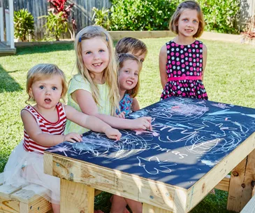 Children sitting around a chalkboard table outdoors, drawing with chalk.