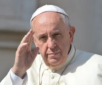 Pope Francis raising his hand, wearing a white cassock and skullcap, with a contemplative expression outside.
