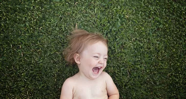 A joyful baby with light brown hair laughing while lying on lush green grass.