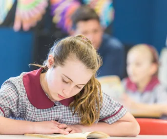 A young girl in school uniform reads a book at a classroom desk, with colorful decorations in the background.