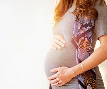 Pregnant woman in a gray dress cradling her belly, with a floral scarf draped over her shoulder.