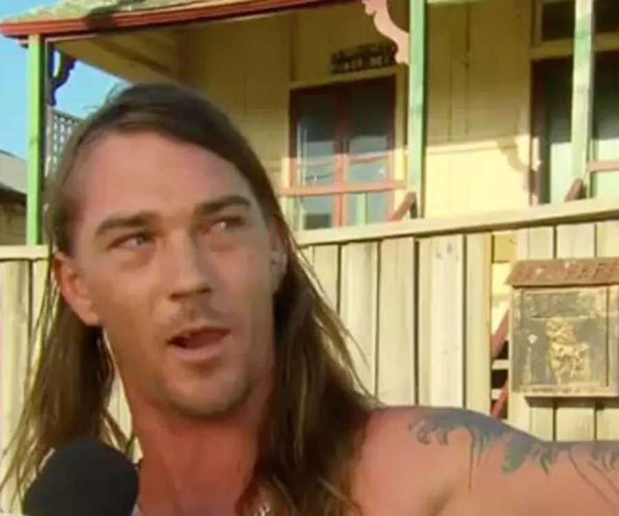 "Man with long hair and tattoos speaking into a microphone outside a wooden fence and house."