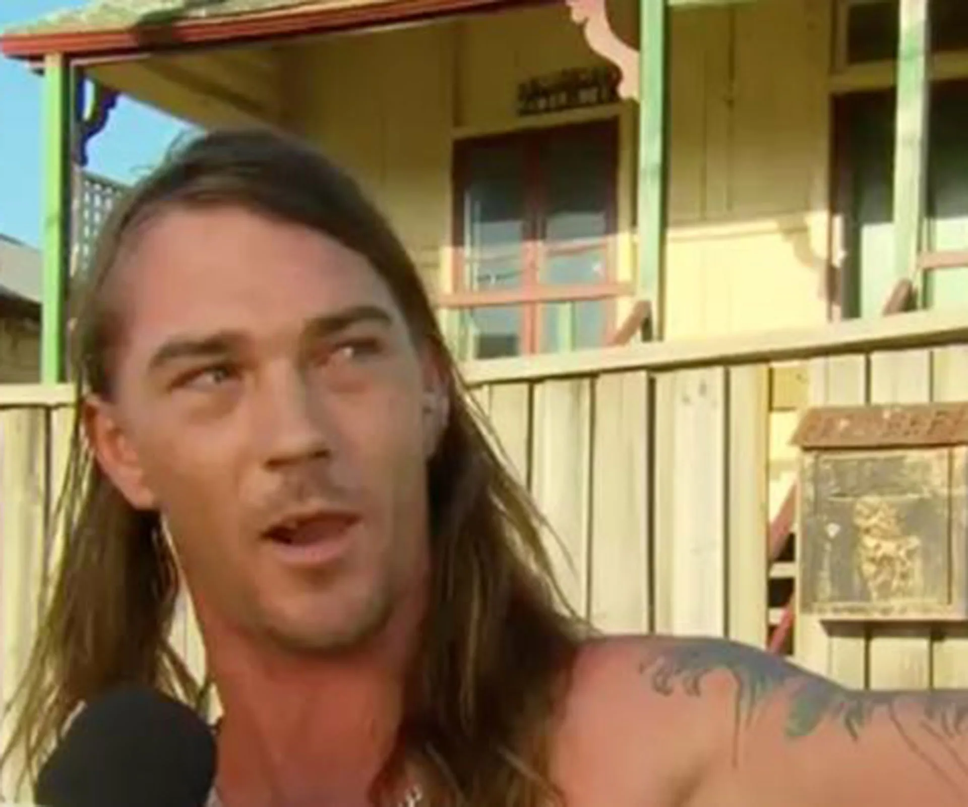 "Man with long hair and tattoos speaking into a microphone outside a wooden fence and house."