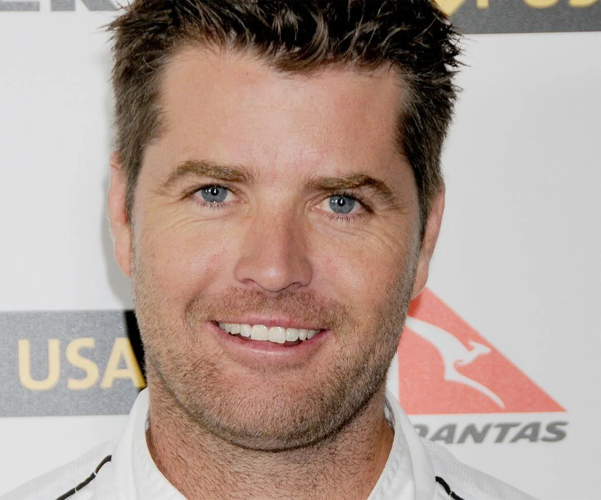 Man smiling at an event with logos in the background.