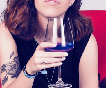 A woman holding a glass of blue wine, wearing a black top, with a floral tattoo on her arm.