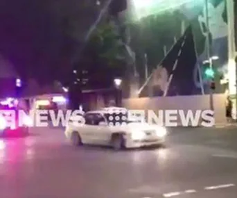 Police car with lights flashing chases a white car at night; street scene with large mural backdrop.