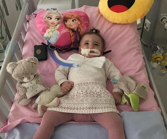 A baby girl lies on a hospital bed surrounded by toys, with a Frozen-themed pillow behind her.