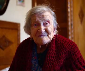 Elderly woman with a gentle smile, draped in a red shawl, seated indoors.