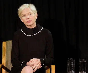 Michelle Williams seated, wearing a black top with white trim, speaking in front of a dark curtain, two water glasses nearby.
