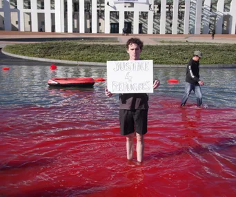 A person stands in red-colored water holding a "Justice 4 Refugees" sign at Parliament House.