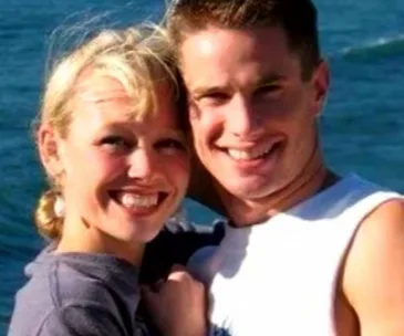 A smiling couple poses closely by the ocean on a sunny day.