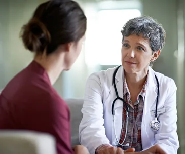 A doctor with a stethoscope talks compassionately with a patient in an office setting.