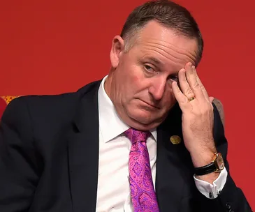 A man in a suit and pink tie touches his forehead, appearing contemplative or stressed, against a red background.