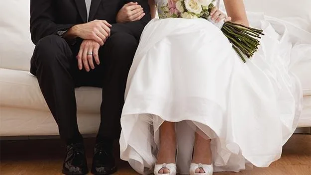 Bride and groom seated, her holding a bouquet, both in formal attire.