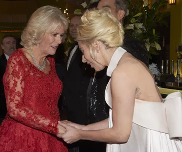 Royal figure in red lace dress warmly greets woman in elegant white gown, holding hands and smiling in a formal setting.