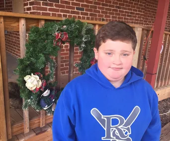 Young boy in blue hoodie stands in front of a festive wreath on a wooden porch with brick wall background.