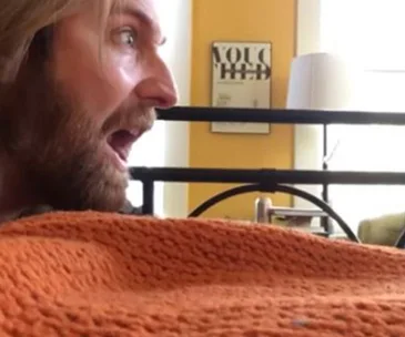 A man with long hair makes a surprised expression at a cat, partially visible behind an orange blanket.