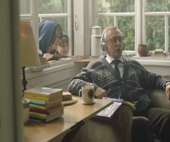 Elderly man with headphones sits by a window with English book; two children peek in smiling.