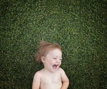 A laughing toddler lying on grass, facing upwards with eyes squinted and mouth open in joy.