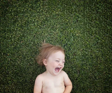 A laughing toddler lying on grass, facing upwards with eyes squinted and mouth open in joy.