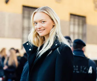 A person with long blonde hair in a black coat smiles outdoors, with blurred figures and buildings in the background.