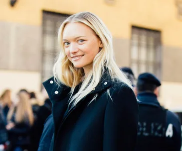 A person with long blonde hair in a black coat smiles outdoors, with blurred figures and buildings in the background.