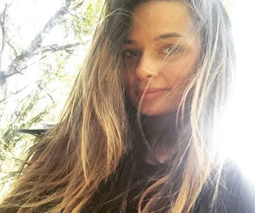 A woman with long hair looks slightly away, with sunlight filtering through trees in the background.