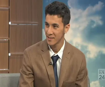 A young man in a suit and tie smiles on a TV set with a cloudy blue background.