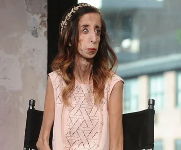 A woman with long hair wearing a headband and a light pink top is seated in a chair, looking thoughtfully to the side.
