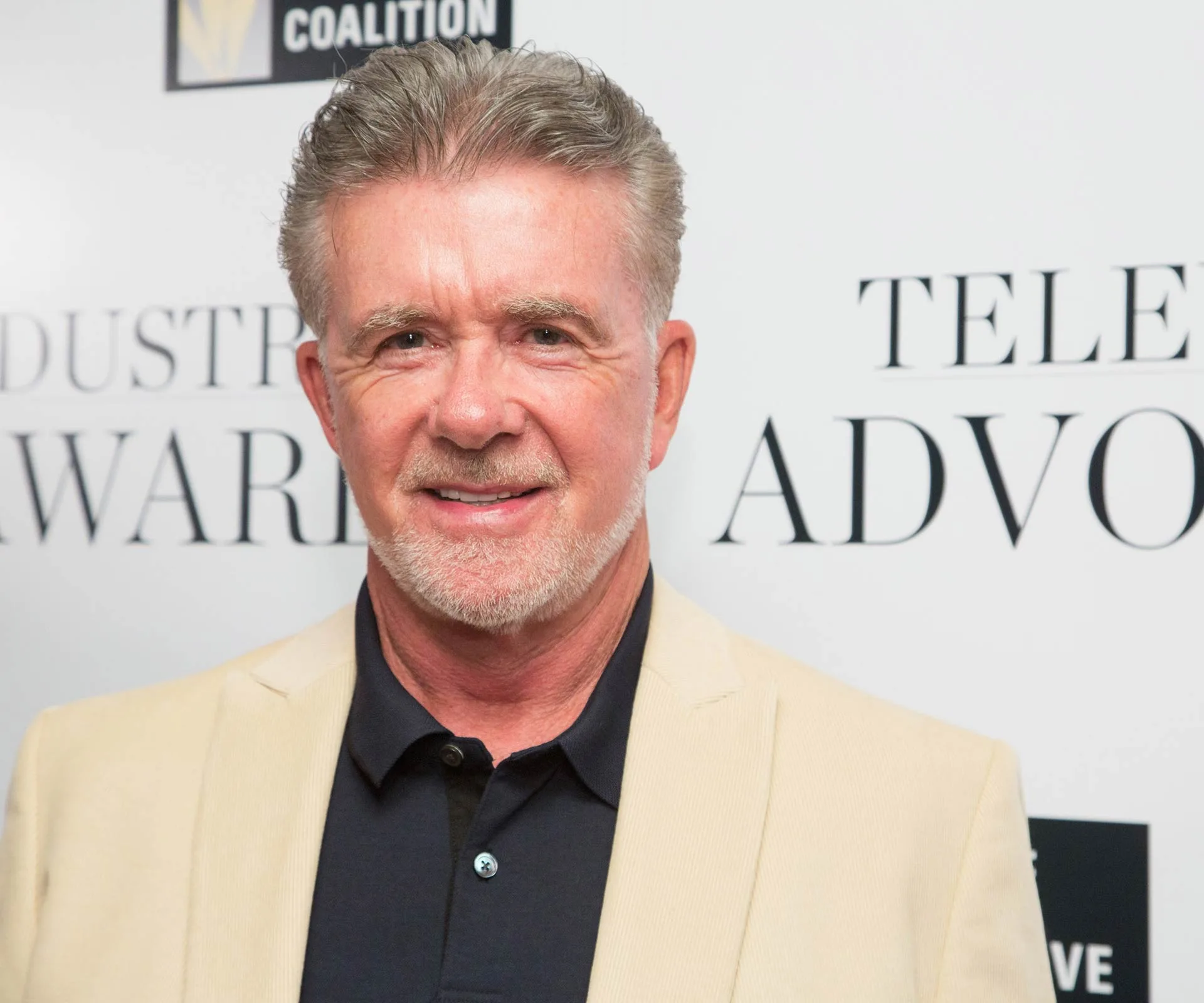 Man in a beige blazer and black shirt smiles at an event backdrop with text in the background.